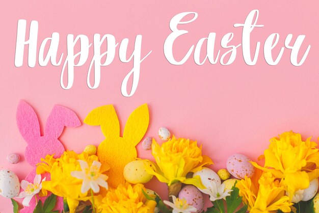 Happy Easter greeting card Happy Easter text and colorful Easter bunnies chocolate eggs and daffodils border on pink background flat lay Seasons greeting card handwritten lettering