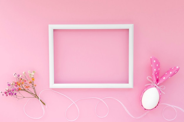 Foto felice concetto di vacanza di pasqua. su sfondo rosa uovo di pasqua con orecchie di coniglio e fiore gypsophila e cornice bianca con spazio vuoto per il testo.