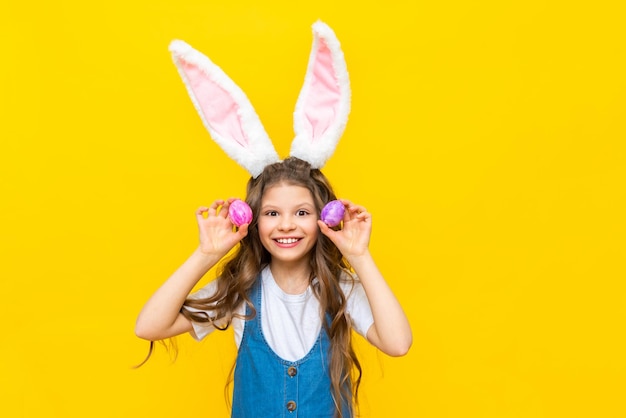 Happy Easter A little girl holds two painted eggs for the holiday A charming baby with rabbit ears on a yellow isolated background