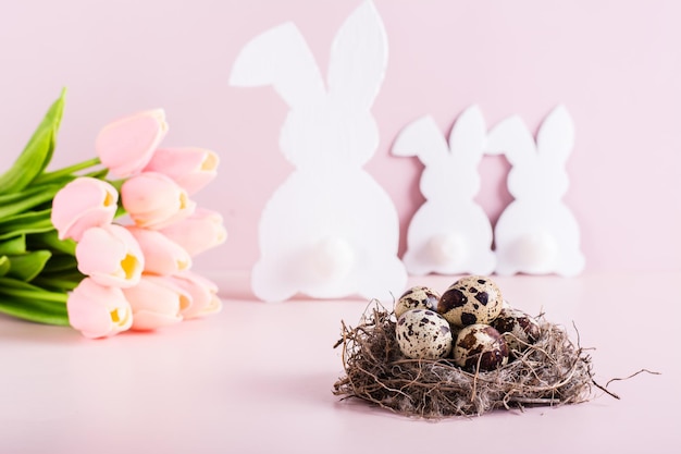 Happy easter nest with quail eggs white rabbit family and bouquet of flowers on pink background