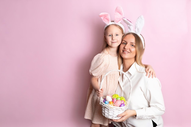 Happy Easter Portrait of beautiful blonde mother and daughter with rabbit ears and a basket of colored eggs in their hands Pink background Space for text High quality photo