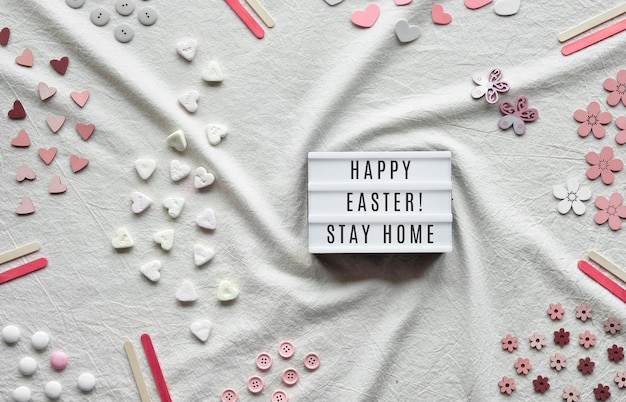 Happy Easter Stay Home text on lightbox Flat lay with light box on ivory off white textile Hearts candy buttons various decor items arranged on cotton tablecloth