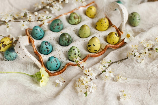 Happy Easter Stylish easter eggs and blooming spring flowers on rustic table Natural painted quail eggs in tray feathers and cherry blossoms on linen fabric Rustic easter still life