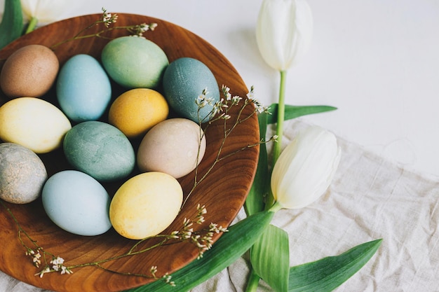 Happy Easter Stylish Easter eggs in wooden plate tulips and linen napkin on rustic white table