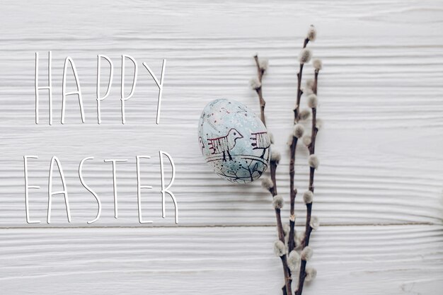 Foto cartolina d'auguri con testo di buona pasqua segno su semplice uovo di pasqua con ornamenti di pulcino e boccioli di salice su sfondo scrivania in legno bianco rustico con spazio per testo vista dall'alto luce soffusa