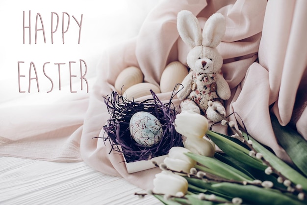 Happy easter text greeting card sign on stylish easter eggs with chick ornaments in nest and bunny rabbit and tulips and willow buds on rustic white wooden background space for text soft light