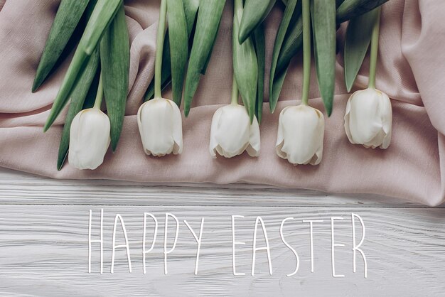 Happy easter text greeting card sign on stylish white tulips on
beige soft fabric and rustic wooden table background top view
atmospheric moment space for text