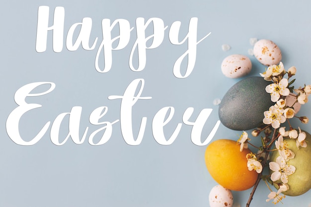 Photo happy easter text and modern stylish blue and yellow easter eggs cherry blossoms flat lay on blue background happy easter greeting card seasons greeting card handwritten lettering