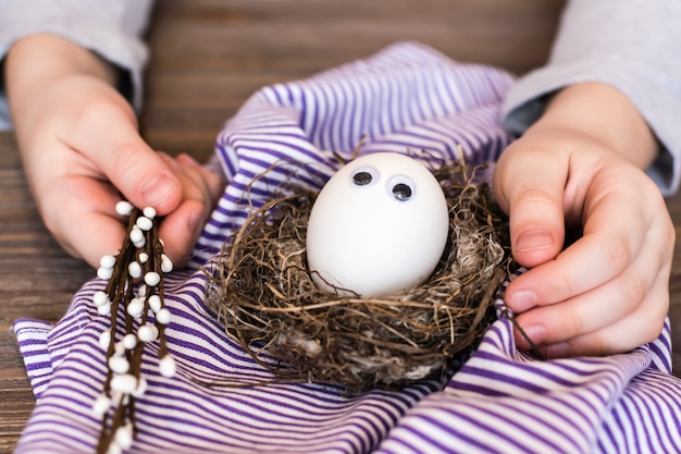 Happy easter. Unpainted egg with eyes in a bird's nest,  children's hands  and willow branches on a wooden table. 