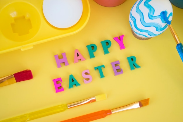 Happy easter word made of colorful letters with eggs in yellow egg tray, paints and brushes paitning egg in blue waves on yellow wall.