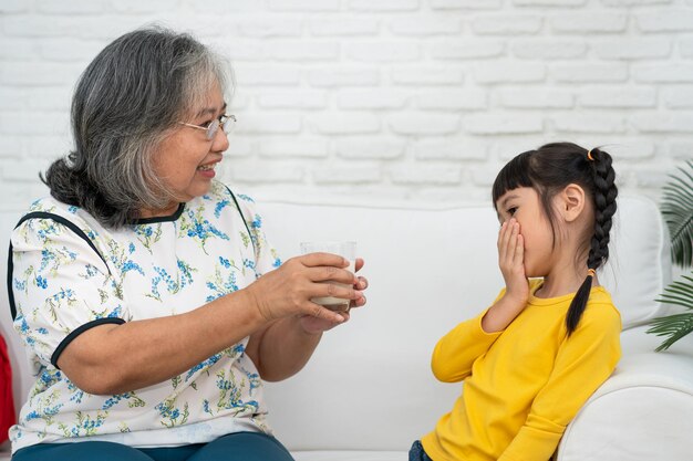 幸せな年配のアジアのおばあちゃんは彼女の孫娘のそばに座って、ガラスから新鮮なミルクを供給します