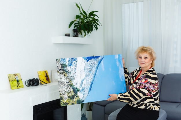 A happy elderly blonde woman is holding a large wall photo canvas at home