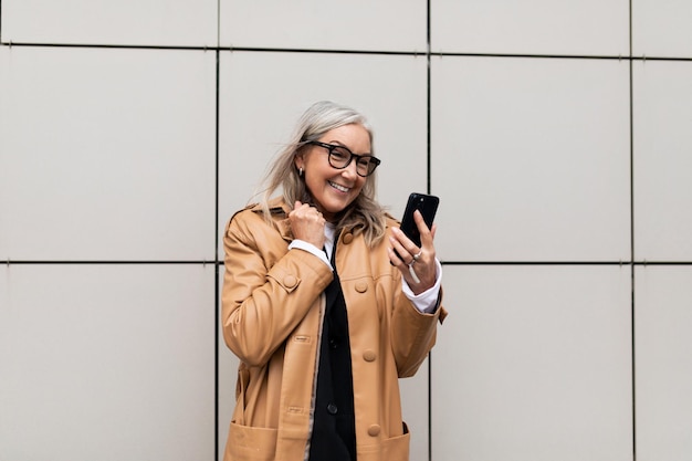 Happy elderly businesswoman with mobile phone rejoices looking at the screen