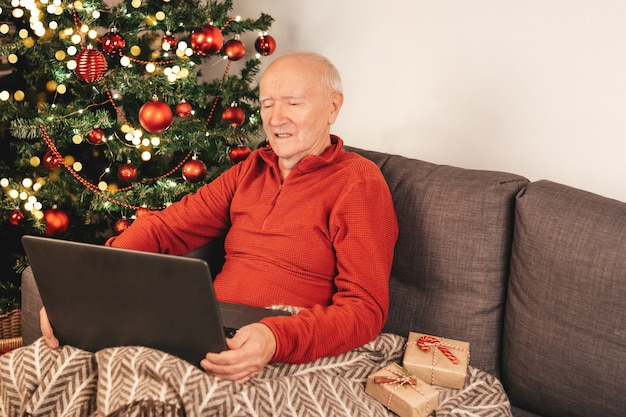 Uomo caucasico anziano felice con il computer portatile che si siede su un divano vicino a un albero di natale con una tazza di tè in chat con i parenti online. autoisolamento, atmosfera vacanziera. scatole regalo.