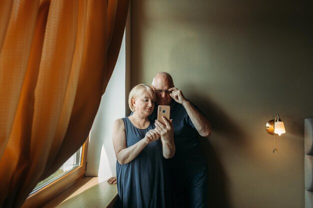 A happy elderly couple stand near the window pose take a selfie on the phone