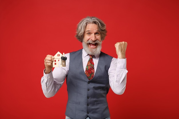 Happy elderly gray-haired mustache bearded man in classic shirt vest tie isolated on red background. People lifestyle concept. Mock up copy space. Holding house and bunch of keys doing winner gesture.