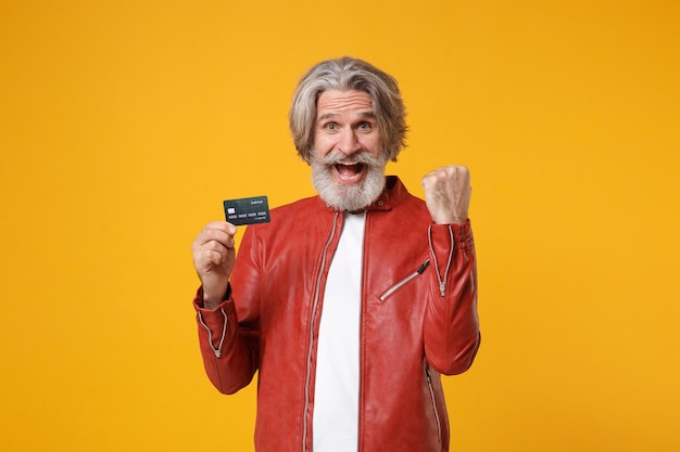 Happy elderly gray-haired mustache bearded man in red leather jacket isolated on yellow orange background. people lifestyle concept. mock up copy space. hold credit bank card, doing winner gesture