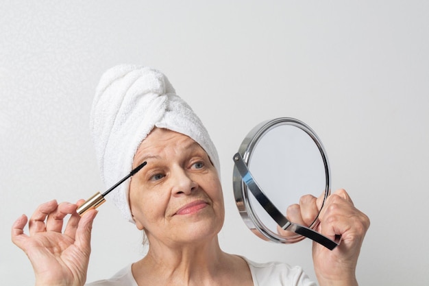 Happy elderly lady with towel on head coloring eyelashes and looking at mirror