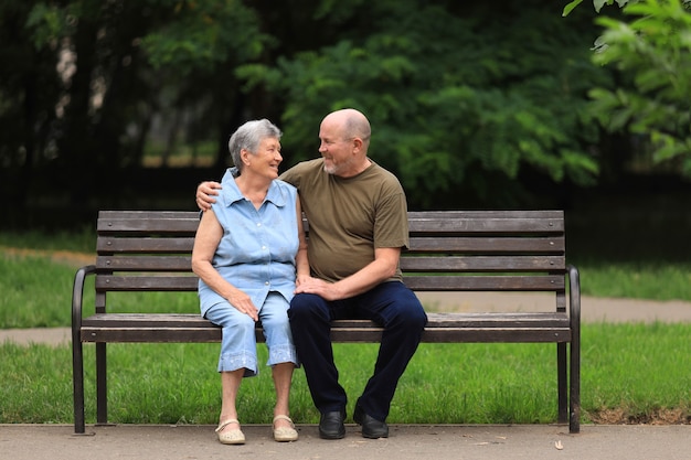 Happy elderly man and disabled woman sit on bench outdoors in summer park