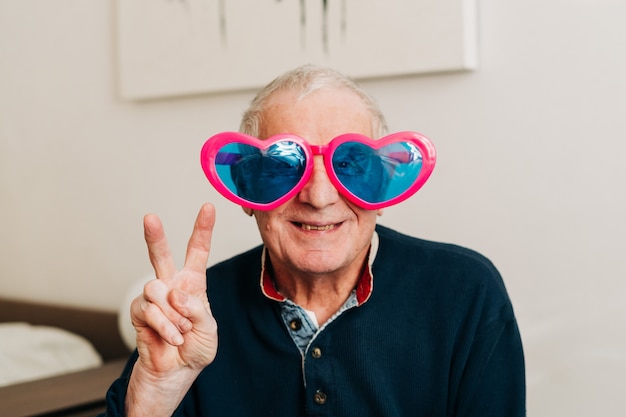 Happy elderly man portrait with heart shaped glasses at home, love concept