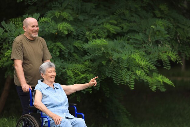 Happy elderly man walking with disabled elderly woman sitting in wheelchair outdoors