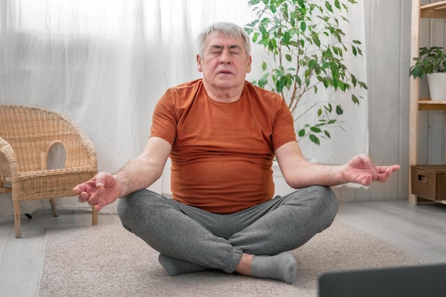 Счастливый пожилой мужчина смотрит онлайн-уроки на ноутбуке. Тренируется дома, сидит на полу в уютной гостиной. Учебные упражнения на пенсии. Старший мужчина занимается йогой в помещении.