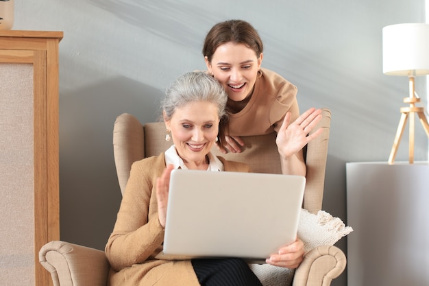 Happy elderly middle mother sitting on chair with her daughter, looking at laptop. Young woman showing video, photos to mommy, trusted relations. Family concept.