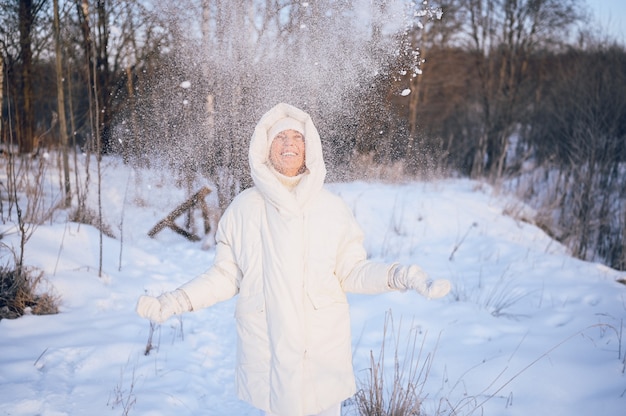 Happy elderly senior mature woman in white warm outwear playing with snow in sunny winter outdoors