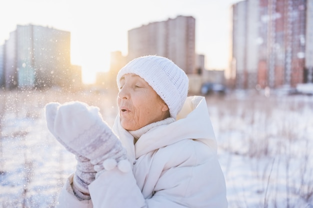 Donna matura senior anziana felice in outwear caldo bianco che gioca con la neve nell'inverno soleggiato all'aperto