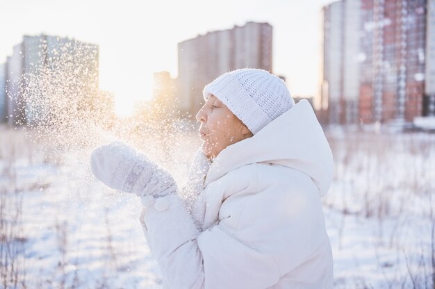 Happy elderly senior mature woman in white warm outwear playing with snow in sunny winter outdoors