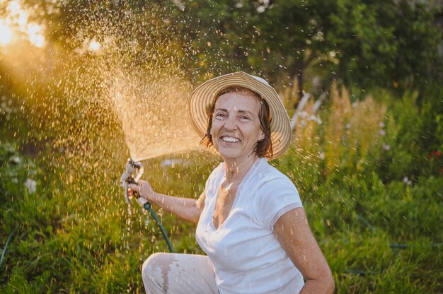 Happy elderly senior woman having fun watering plants with hose in summer garden drops of water in