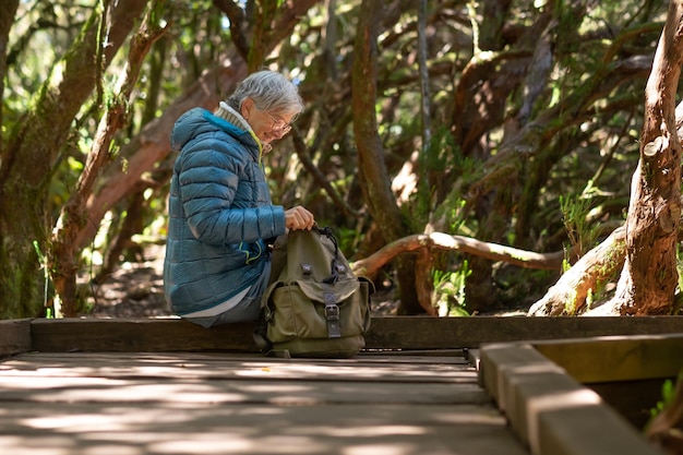 Happy elderly traveler woman enjoying hike in nature sitting in\
the woods looking into her backpack