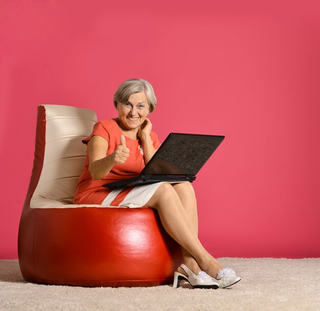 Happy elderly woman sitting with laptop on red armchair