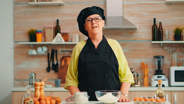 Felice donna anziana che indossa bonete guardando la fotocamera nella sala da pranzo a casa. vecchio fornaio in pensione con uniforme da cucina che prepara ingredienti da forno sul tavolo pronto per cucinare pane, torte e pasta fatti in casa.