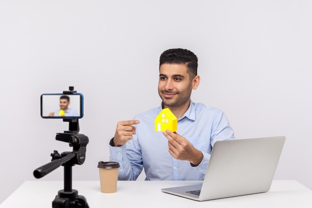 Happy elegant man real estate agent sitting in office workplace and showing paper house to camera recording video streaming online blog about purchase or rental housing indoor studio shot isolated