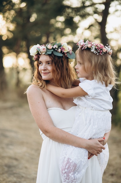 Foto felice elegante madre e sua figlia bella con ghirlande di fiori in un parco al tramonto