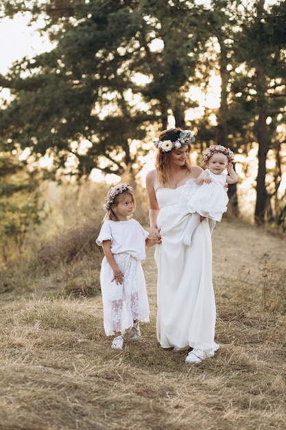 Happy elegant mother and her two beautiful daughters with flower wreaths in nature at sunset