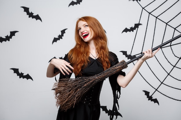 Счастливый элегантный ведьма пользуются играть с метлой Хэллоуин партии на сером фоне.
