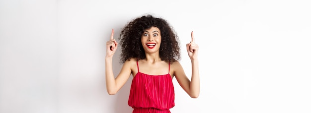 Photo happy elegant woman in red dress and makeup smiling amused and pointing fingers up at logo showing a