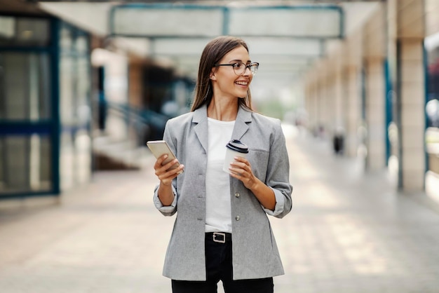 A happy elegant young businesswoman standing in the business center holding takeaway coffee