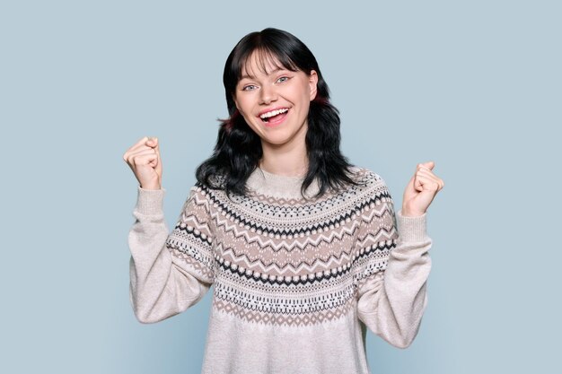 Happy emotional young woman showing gesture clenching fists like winner raising hands up on grey color studio background Wintertime beauty lifestyle holiday New Year Christmas concept