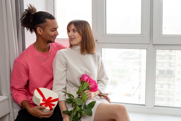 Happy enamored guy in a pink jumper and a girl in a white dress sit by the window