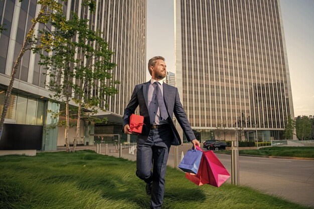 オフィスのメンズデーの外で買い物袋とギフトボックスを保持しているスーツの幸せな起業家