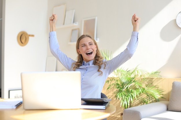 Happy entrepreneur woman sit at desk reading good news and express joy rising hands up.