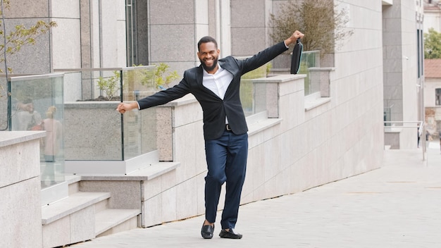 Happy ethnic African American man worker businessman bearded manager leader entrepreneur executive employer in city dancing happy funny walking street cheerful with new job pretend flying airplane