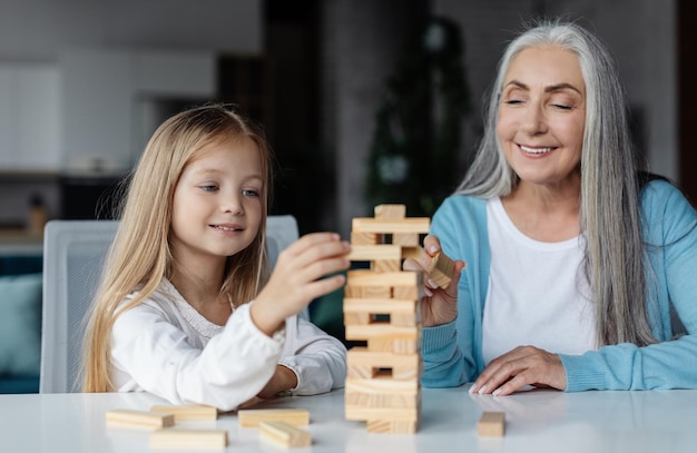 Happy european little girl and old woman play in tower game on table in living room interior