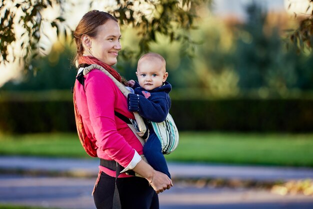 Happy european woman with little boy in sling walks in park