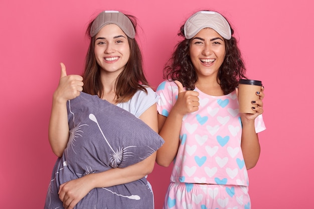 Happy european women standing with satisfied facial expressions, having fun together, posing in sleeping mask and pajamas, holding coffee in disposable cup, show thumb up, wake up with good mood.