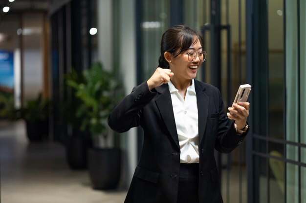 写真 幸せな興奮 成功 良い知らせ 笑顔のある若いアジア人女性の肖像画 携帯電話を使う幸せなアジア人ビジネスウーマン 現代的なオフィスワークプレイス