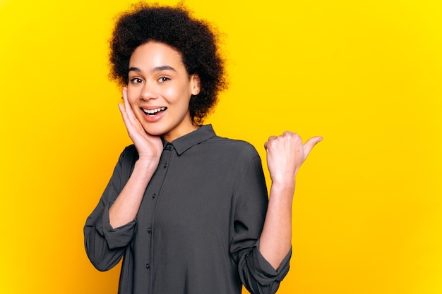 Photo happy excited african american or brazilian young woman with short curly hair looking at camera points finger to the side behind the back to empty space stand on isolated yellow background smiling